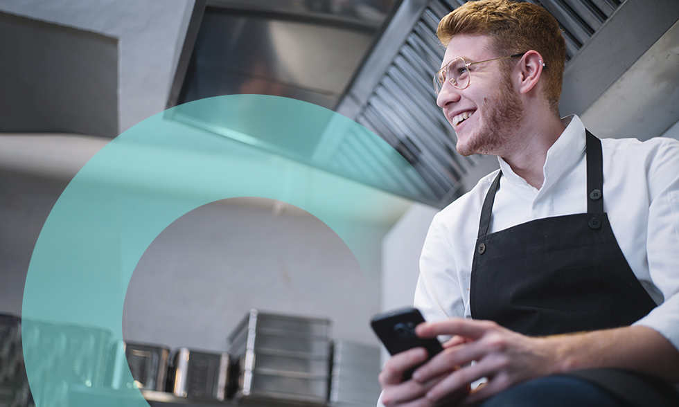 Side view of young man in cook uniform leaning on kitchen counter and using modern smartphone while standing in restaurant kitchen
